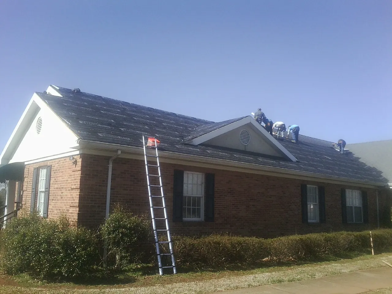 Crew tearing shingles off a brick ranch roof with a ladder set — Metal Roof Repair work by our crew in Hopwood