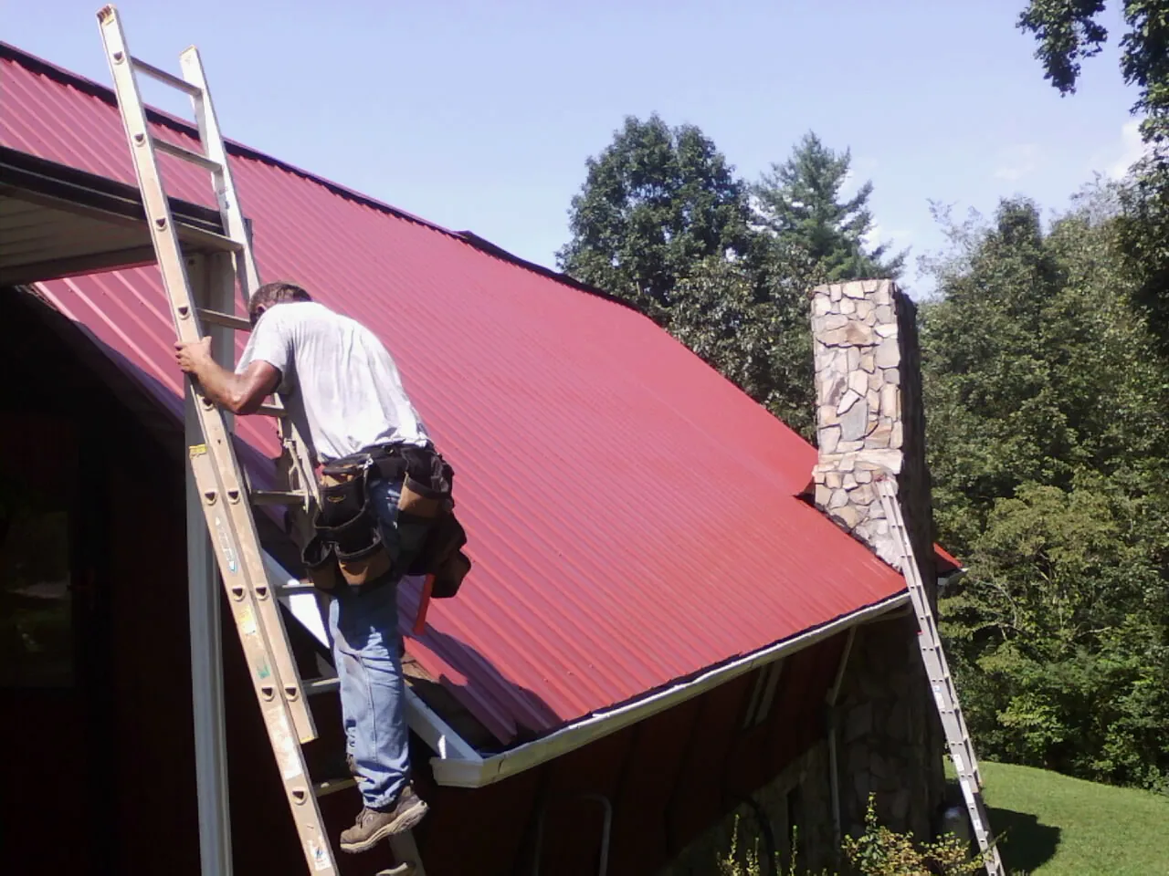 Roofer on a ladder beside a red metal roof and stone chimney — Soffit & Fascia Repair by Roofing Brothers in Hopwood